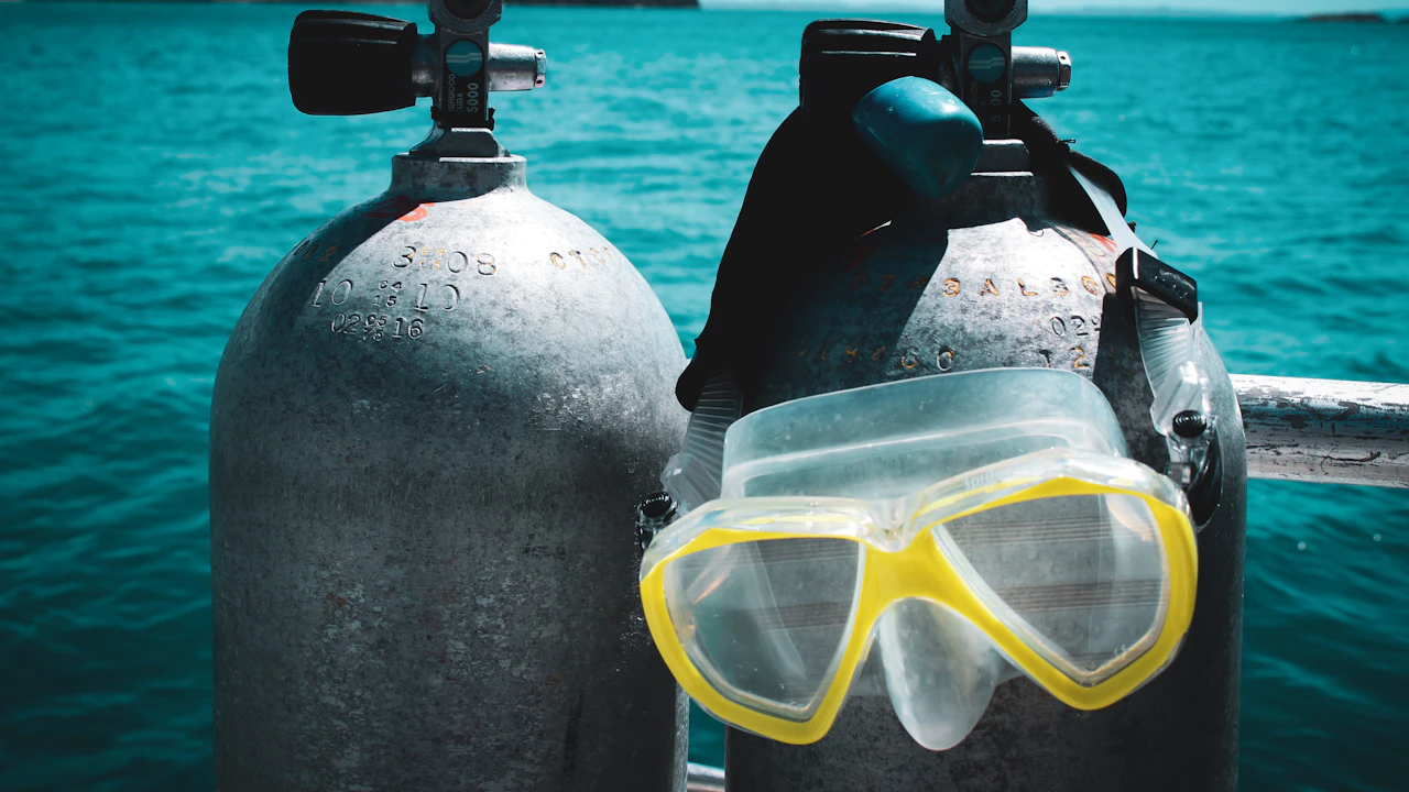 Two scuba tanks and a yellow dive mask on a boat rail above turquoise water