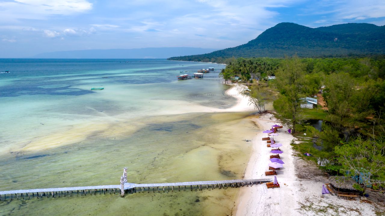 Aerial view of a long wooden pier over turquoise water beside a palm-lined beach