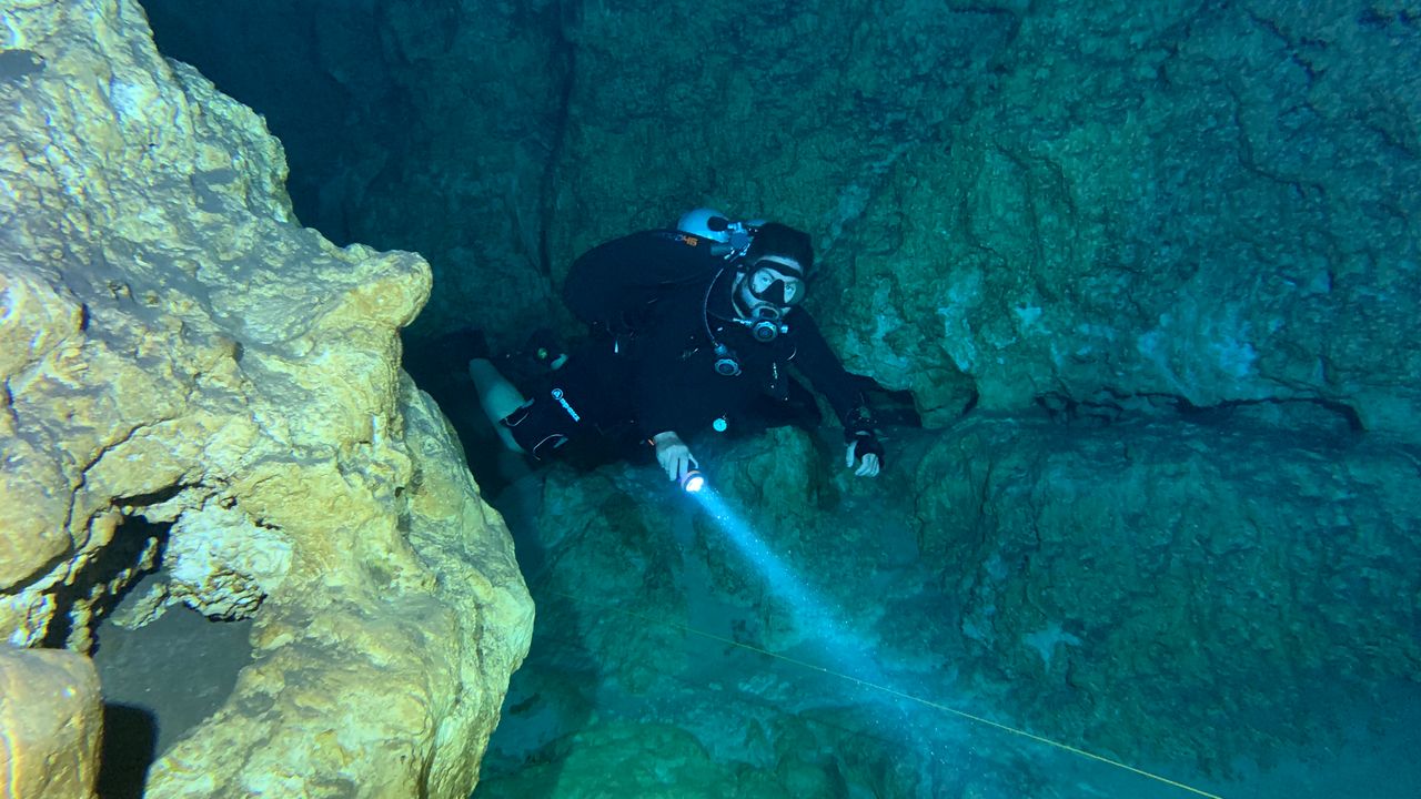 Diver with a torch swimming past hanging limestone formations in dim blue water