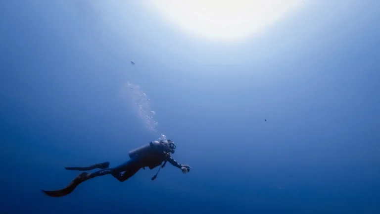 Scuba diver swimming alone through deep blue water below a bright sun