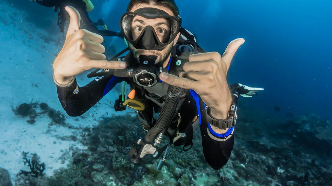 Scuba diver giving a hand signal toward the camera above a reef, another diver behind
