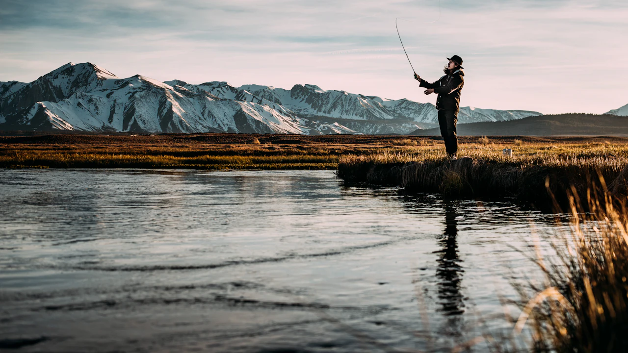 Angler casting a fly line on a calm river below snowy mountains