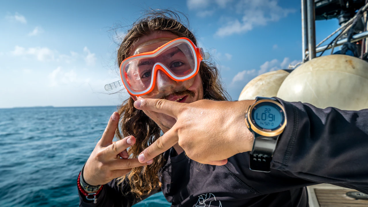 Smiling diver in a pink mask pointing at a dive computer on a boat