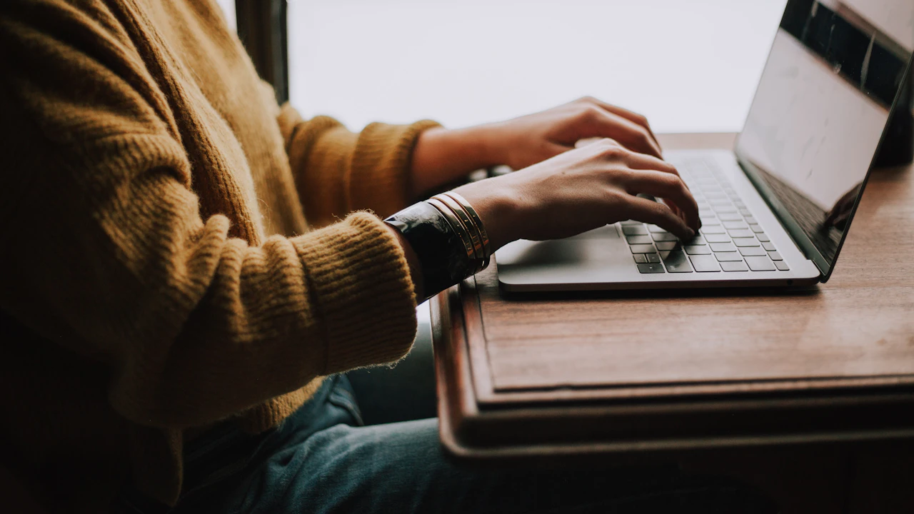 Person in a mustard sweater typing on a laptop at a wooden table by a window