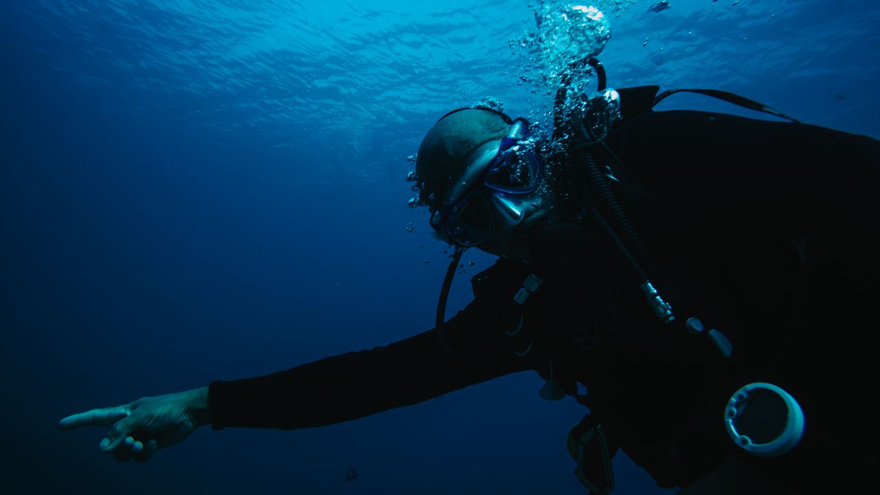 Scuba diver in a dark wetsuit reaching forward while swimming in deep blue water