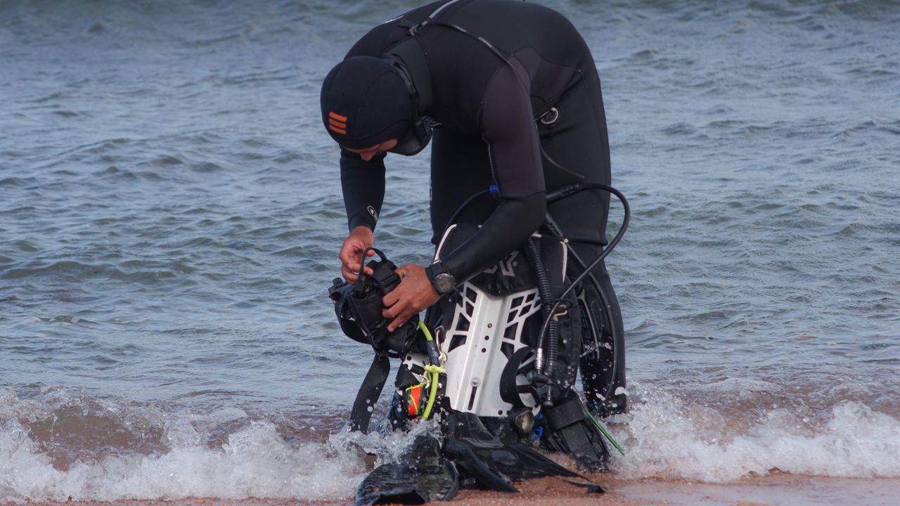 Diver in a wetsuit handling scuba gear in the shallow surf